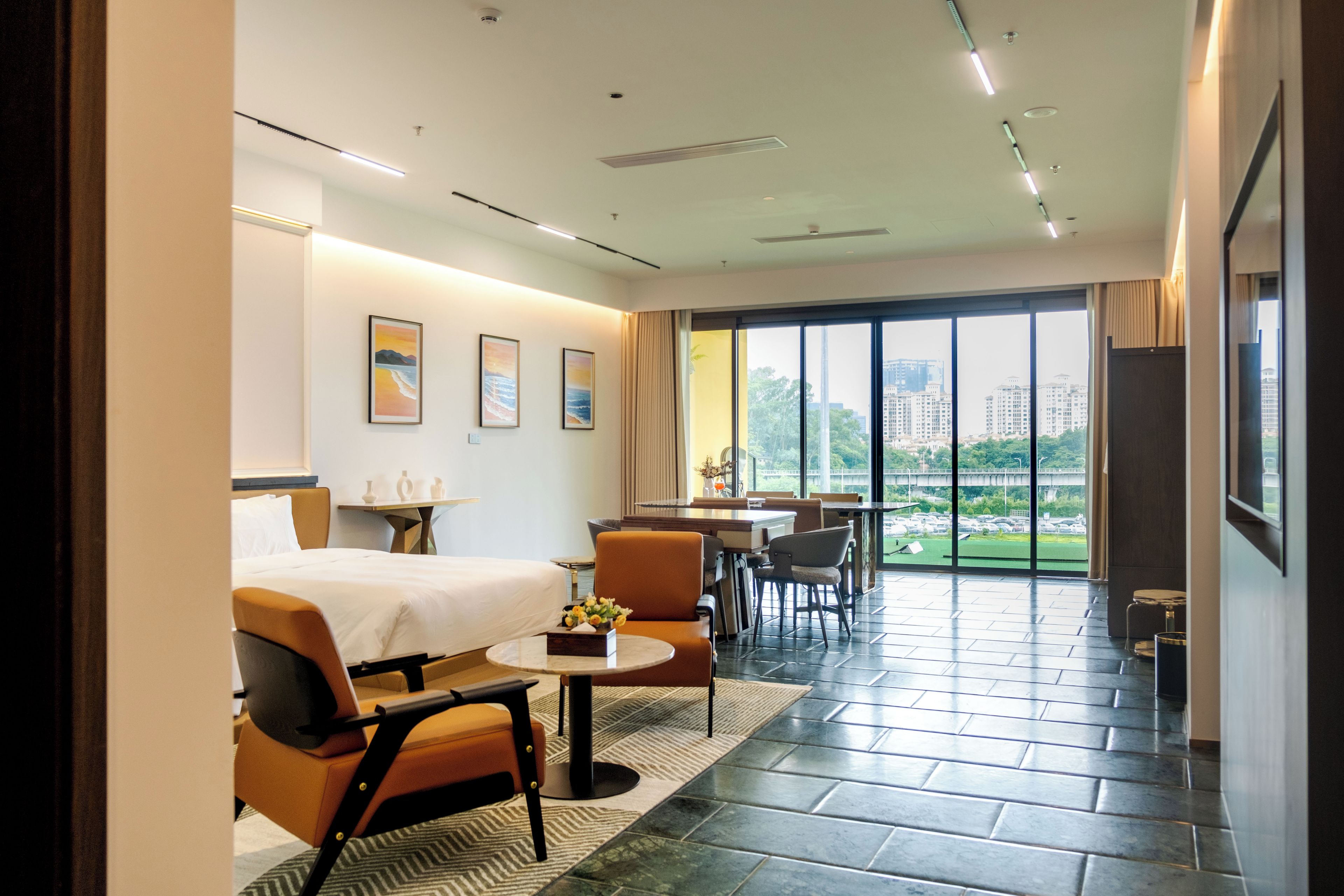 A hotel room with a bed, a dining table, chairs, a small table with a vase, and a large window with a view of buildings.