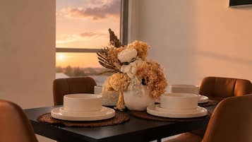 A dining table set with white plates and bowls, a centerpiece of dried flowers and greenery, and a view of the sunset through a window.