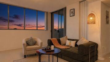 Ein Wohnzimmer mit Sofa, Sessel und Couchtisch. Ein großes Fenster bietet Blick auf den Himmel.