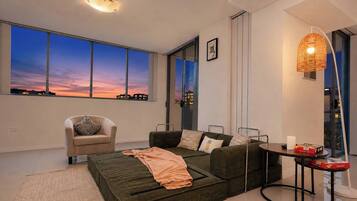 Un salon moderne avec un canapé, un fauteuil et une table à manger. De grandes fenêtres offrent une vue sur le paysage urbain au crépuscule.