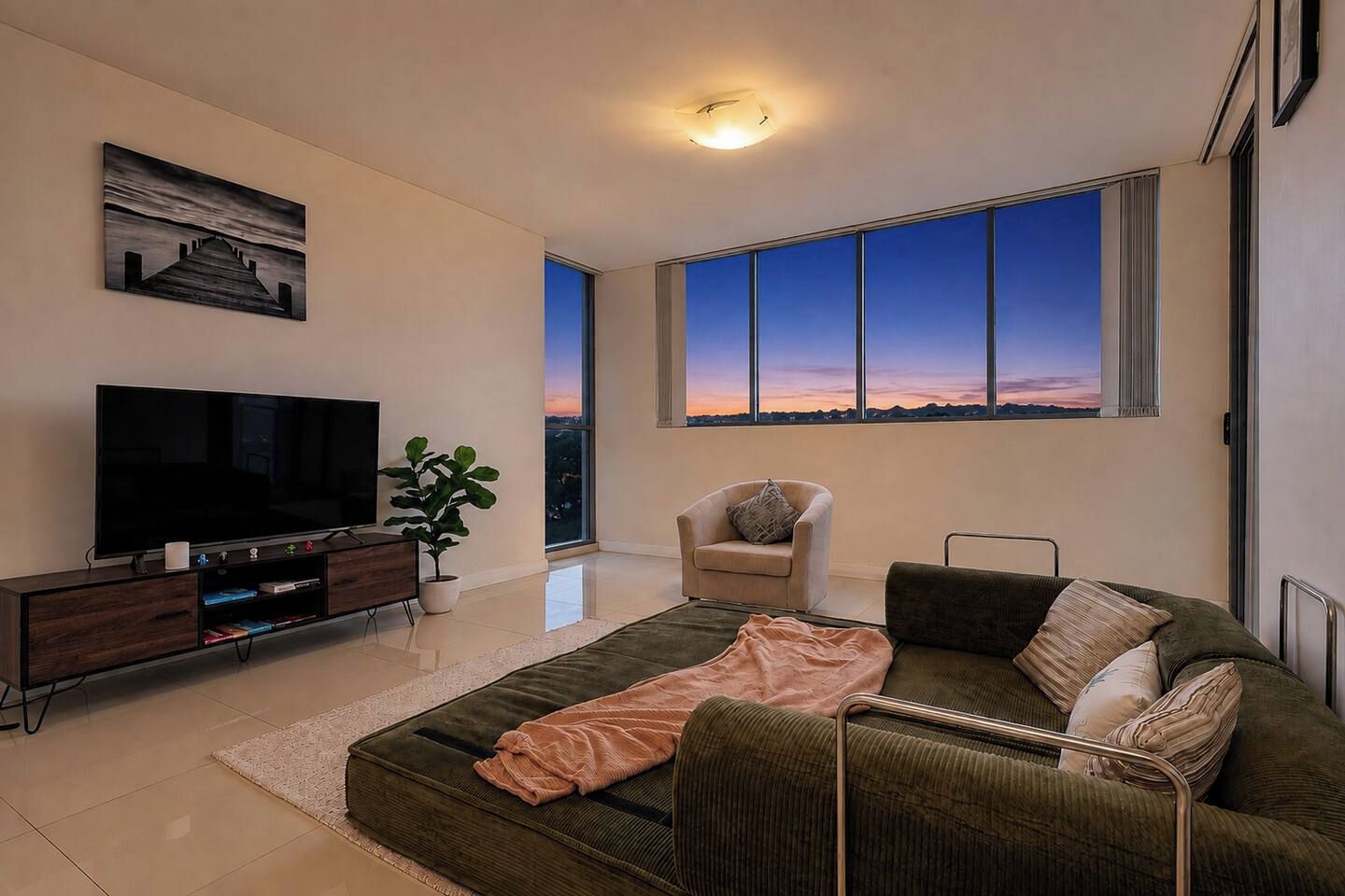 A modern living room with a large flat-screen TV, a sofa, a chair, and a large window offering a view of the sunset.