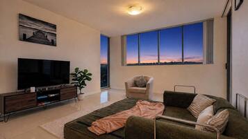 A modern living room with a large flat-screen TV, a sofa, a chair, and a large window offering a view of the sunset.