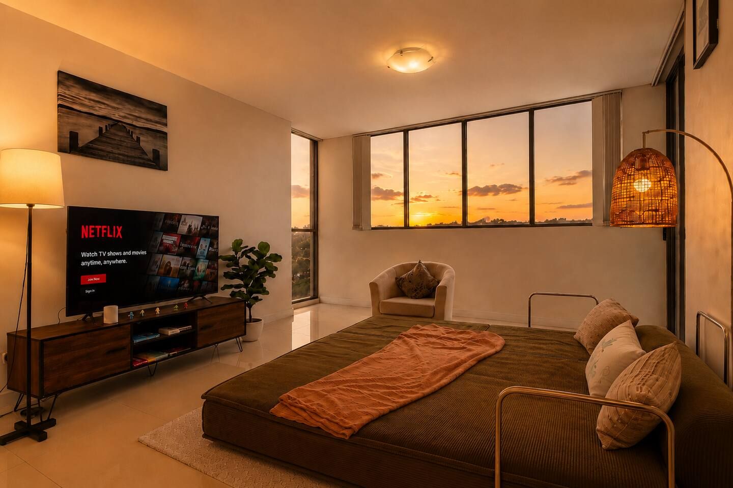 A modern bedroom with a large bed, a TV, a chair, and a view of the sunset through large windows.