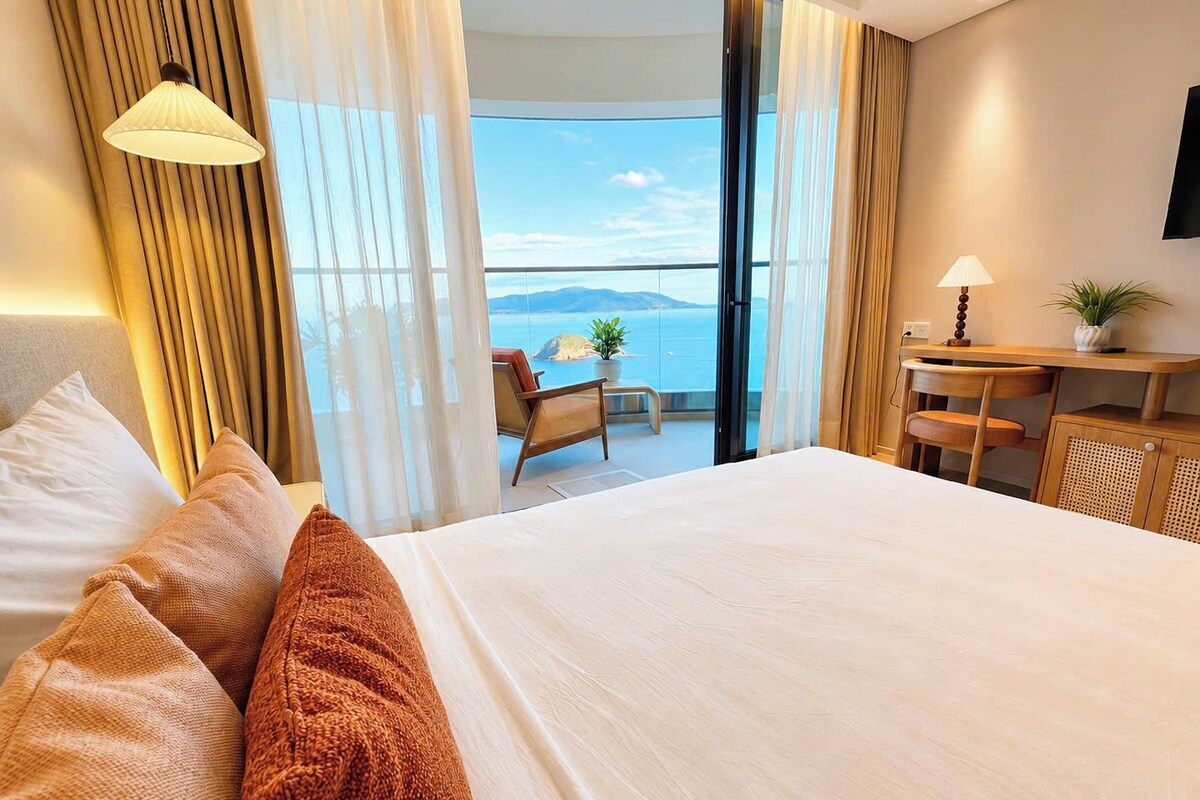 Quarto de hotel com uma cama grande, escrivaninha, cadeira e vista para o mar.