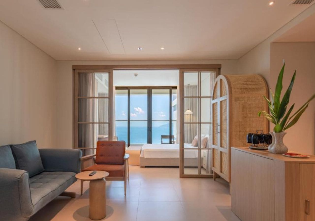 Une chambre d’hôtel moderne, dotée d’un canapé, d’un fauteuil, d’une table de chevet et offrant une vue sur la mer grâce à de grandes fenêtres.