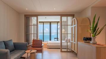 Une chambre d’hôtel moderne, dotée d’un canapé, d’un fauteuil, d’une table de chevet et offrant une vue sur la mer grâce à de grandes fenêtres.