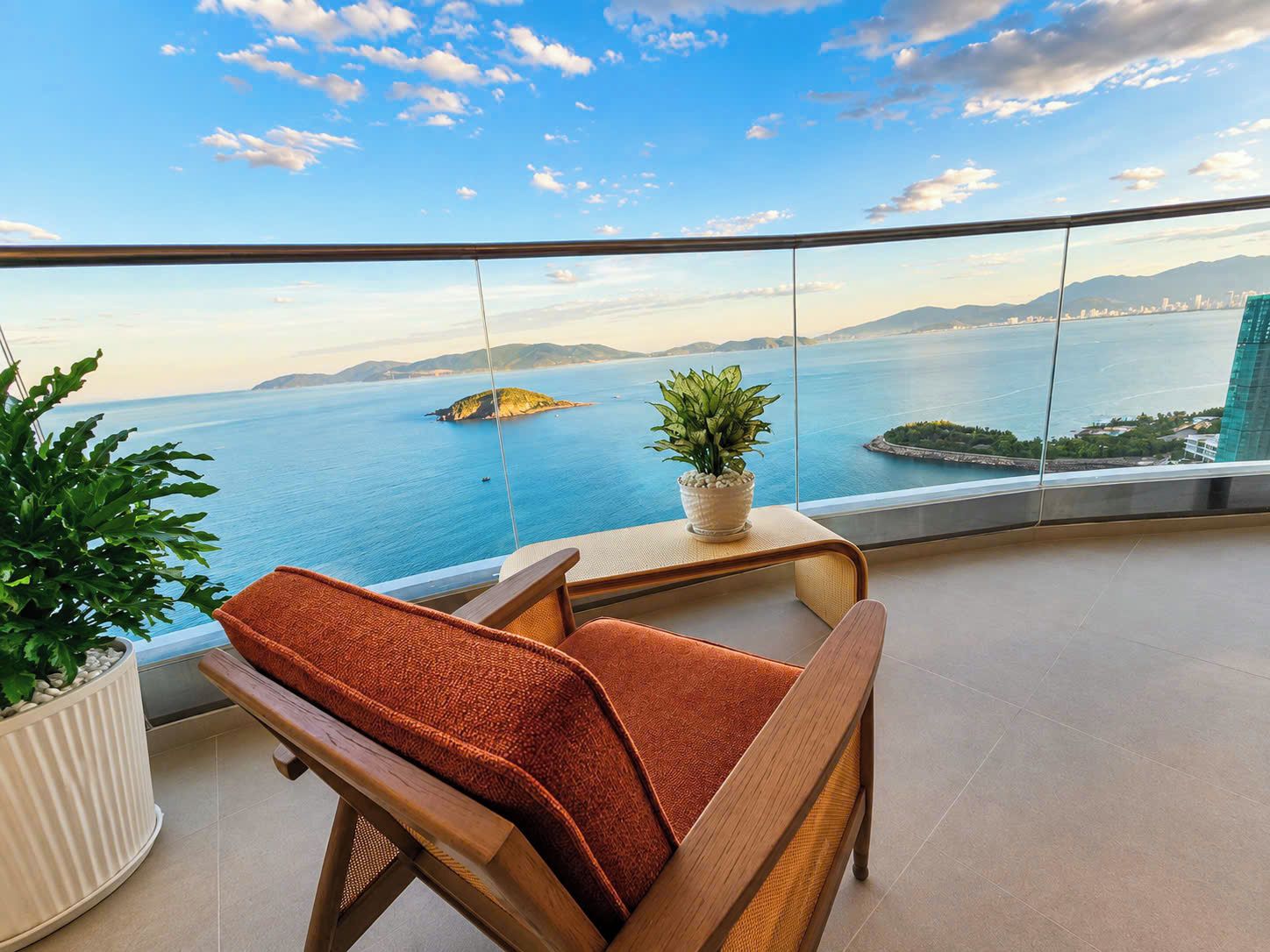 Ein Balkon mit Blick auf das Meer und ferne Berge, ein Holzstuhl, ein Blumentopf und eine weiße Heizung.