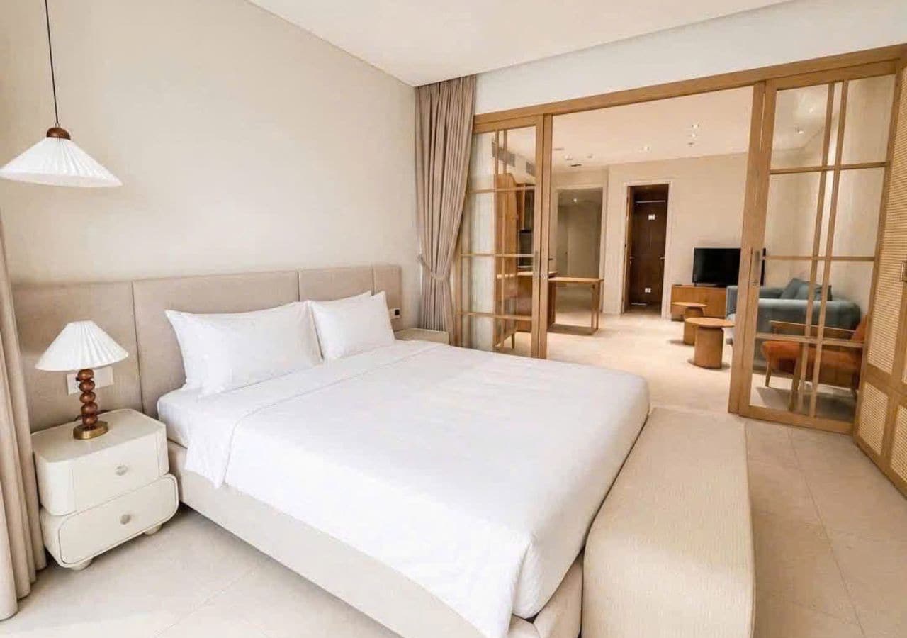 A modern hotel room with a large bed, a nightstand with a lamp, and a sliding glass door leading to a sitting area.