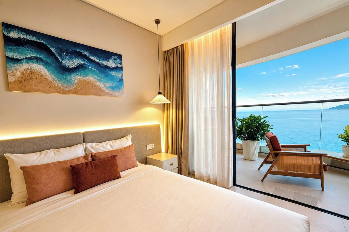 Ein Hotelzimmer mit einem großen Bett, einem Nachttisch, einer Lampe, einem Gemälde mit einer Strandansicht und einem Balkon mit Meerblick.