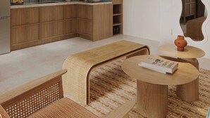 Cozinha moderna com armários de madeira e uma ilha integrada, uma área de estar com banco de fibra natural e uma mesa redonda com um livro.