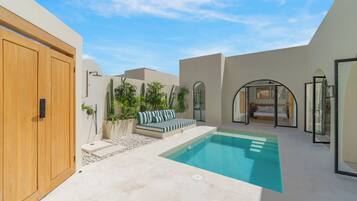 A modern outdoor pool area with a wooden door, a seating area with striped cushions, and a bedroom visible through an arched window.