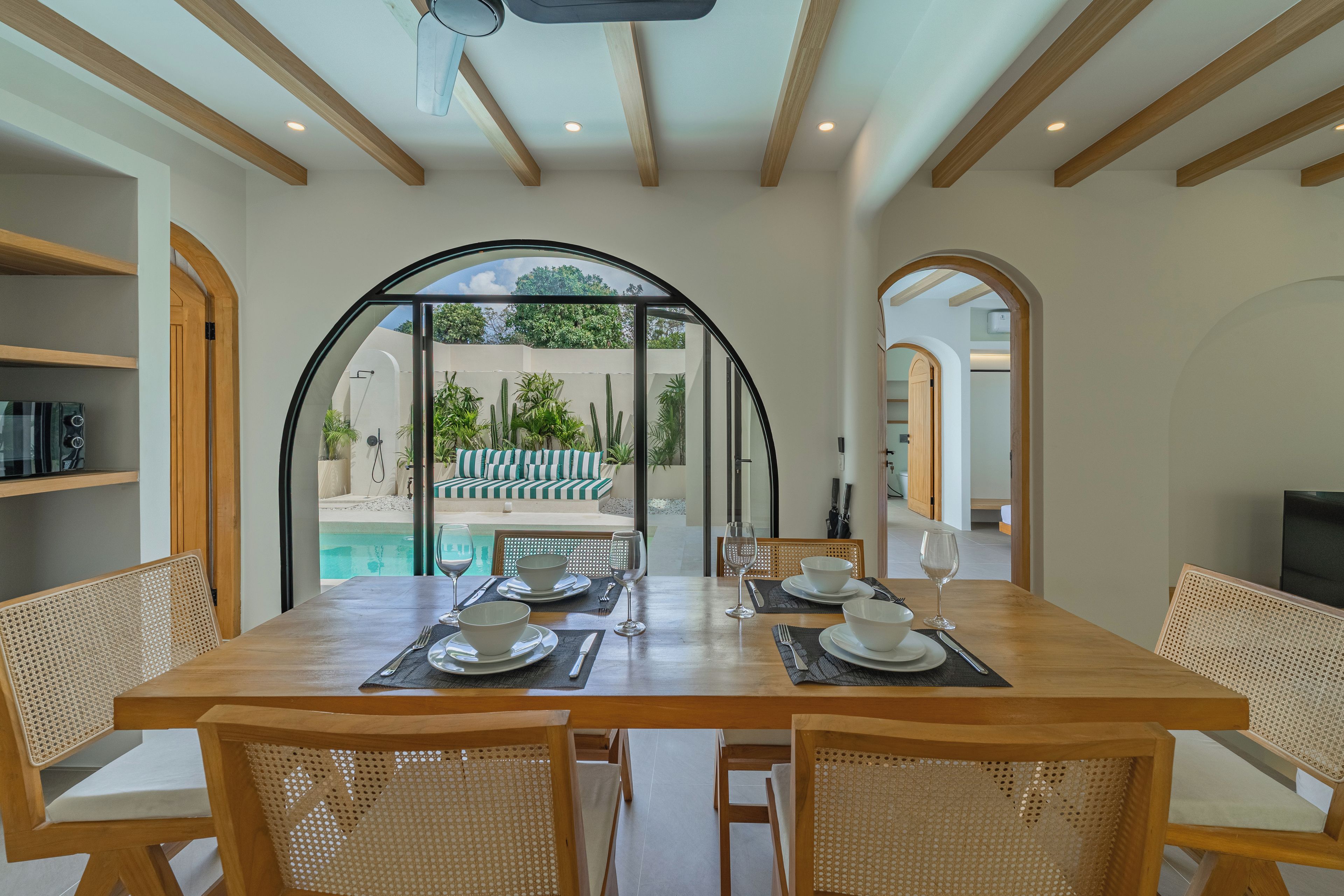 A dining area with a view of a pool and garden, wooden beams on the ceiling, and a glass door leading outside.