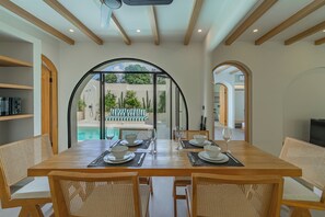 A dining area with a view of a pool and garden, wooden beams on the ceiling, and a glass door leading outside. - Villa Solmea Bingin (Bingin)