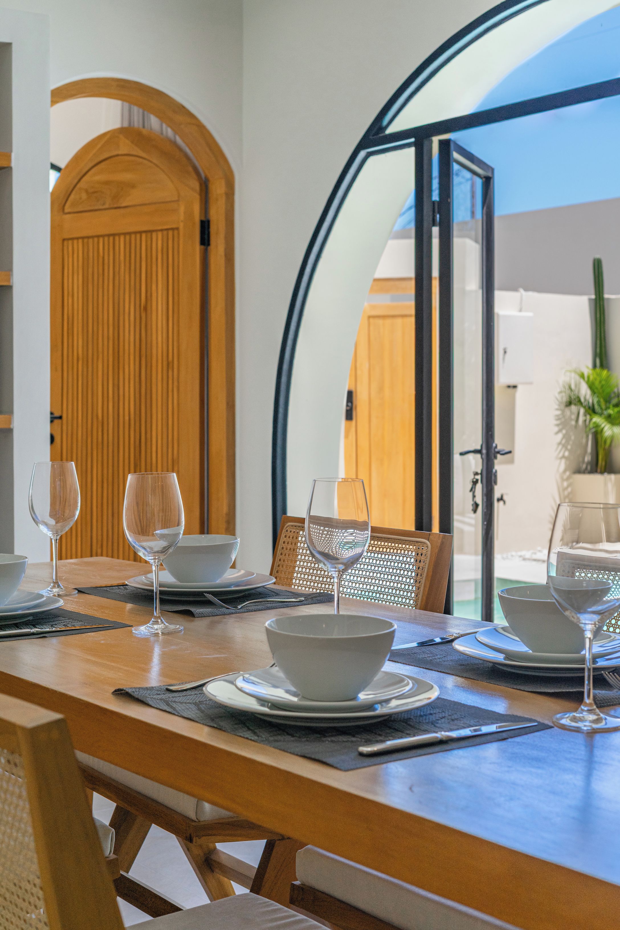 A dining table set with white bowls and wine glasses, a wooden chair, and a view through a glass door to the outside.