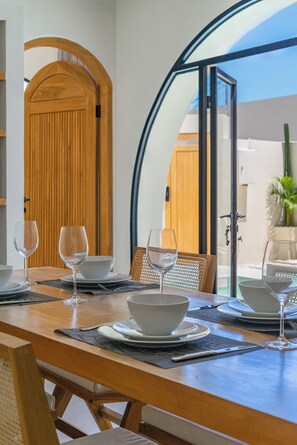A dining table set with white bowls and wine glasses, a wooden chair, and a view through a glass door to the outside. - Villa Solmea Bingin (Bingin)