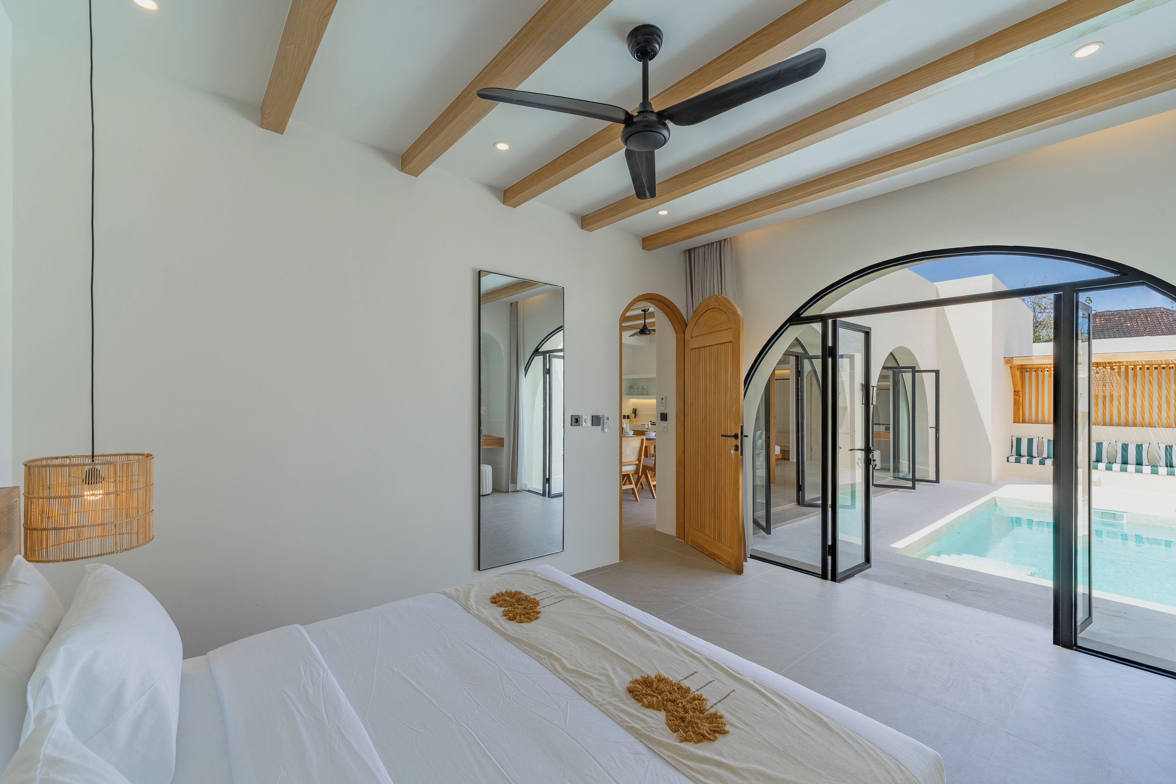 Un dormitorio con cama, ventilador de techo, un espejo grande y vista a una piscina.