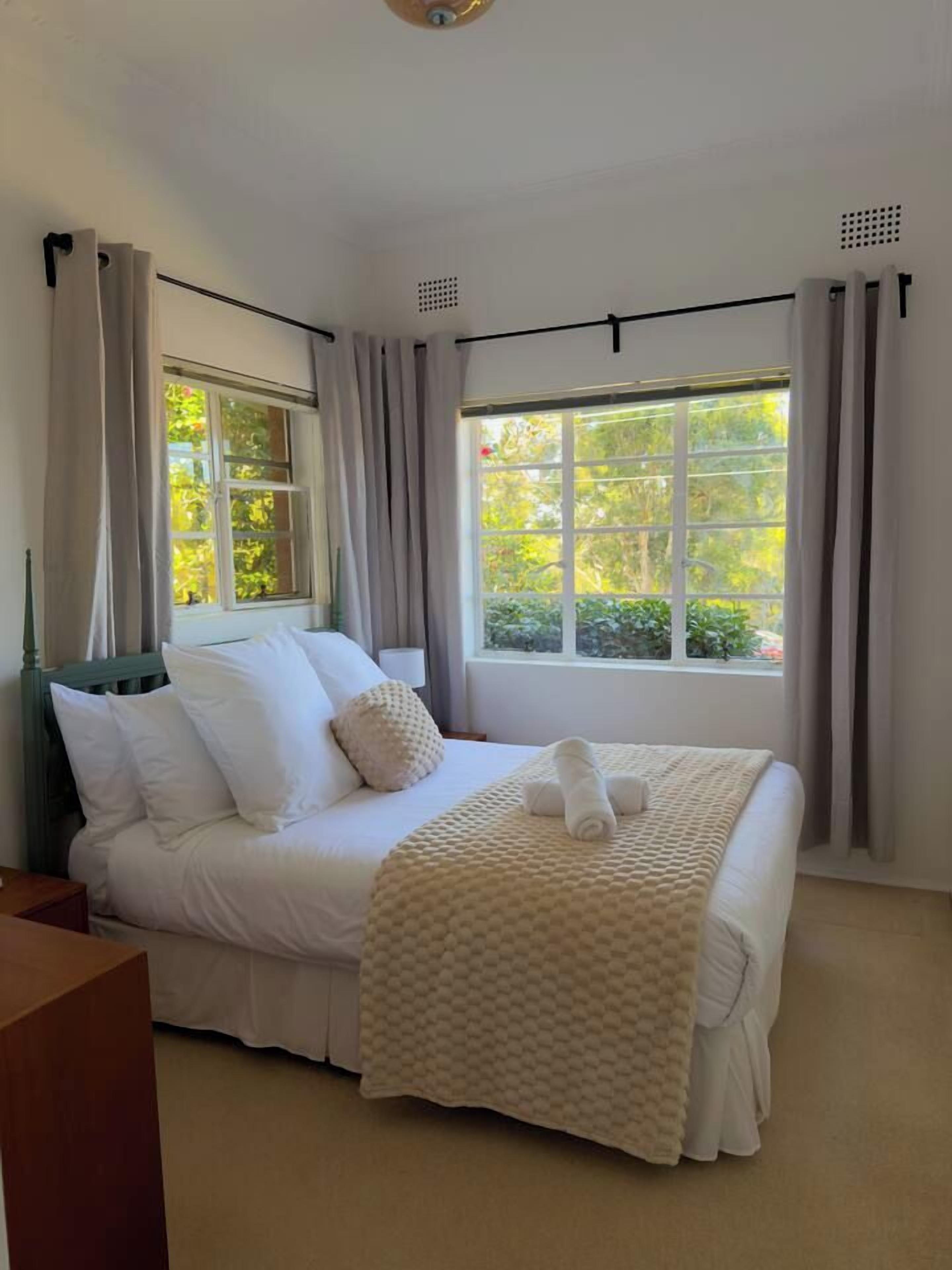 A neatly made bed with white linens and a beige throw blanket, positioned in a room with large windows and curtains.