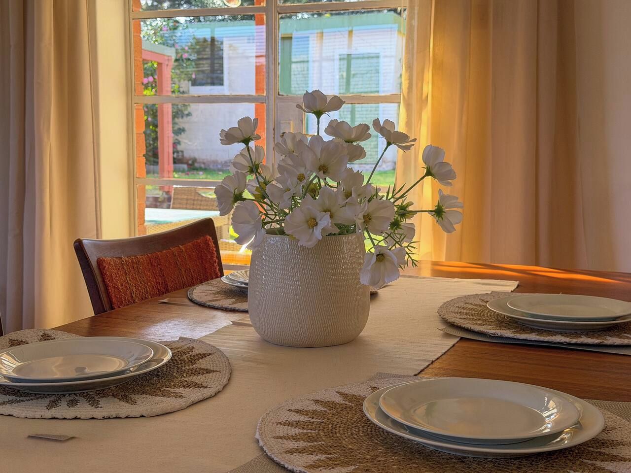 A dining table set with plates, a vase with white flowers, and a view through a window.