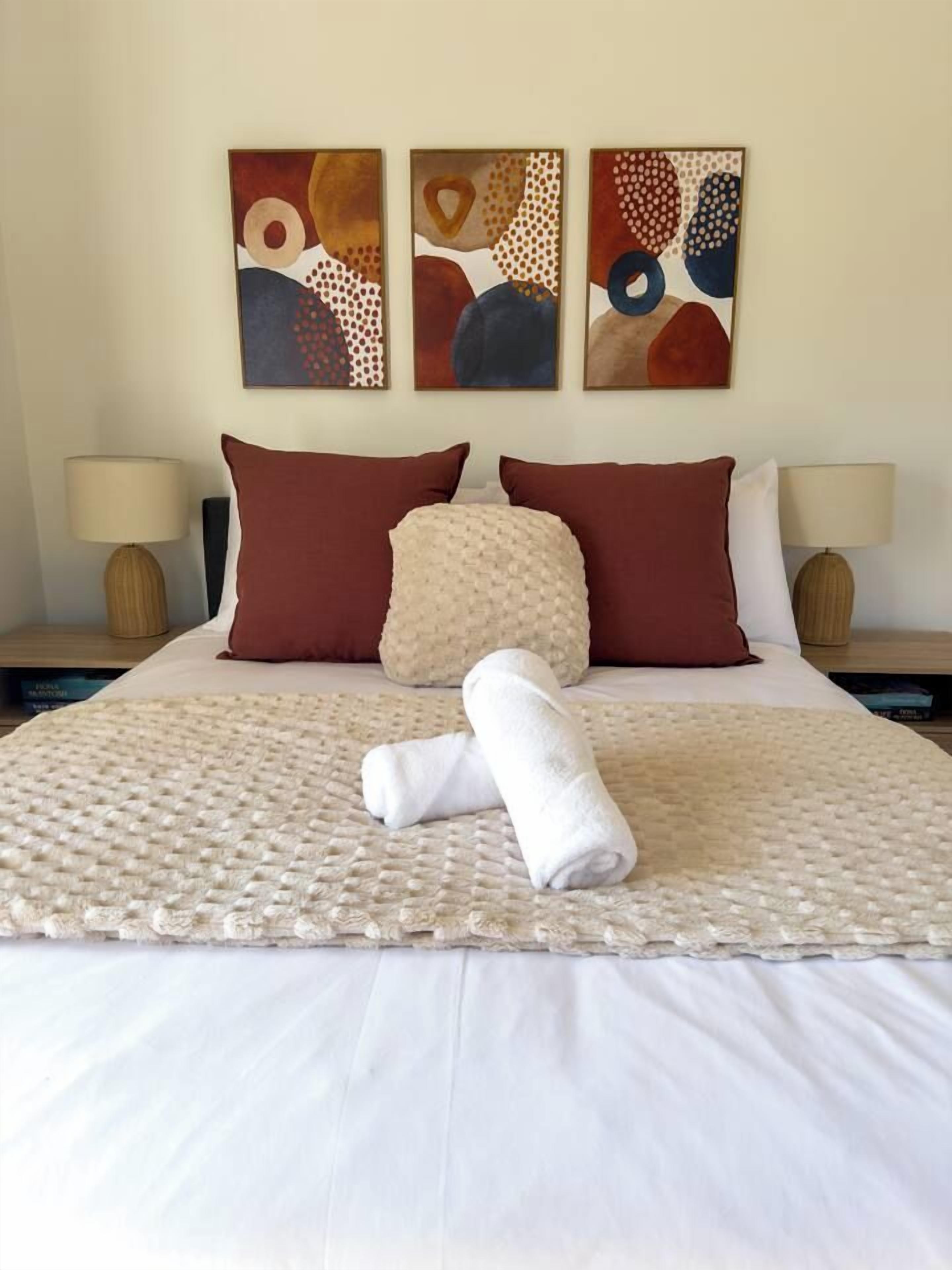 A neatly made bed with a beige blanket and two pillows, a rolled towel, and abstract wall art above.