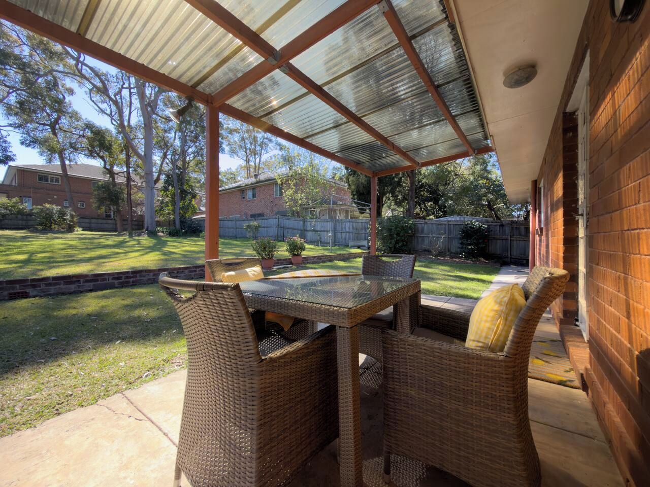 A covered patio with wicker furniture and a glass table, overlooking a backyard with a lawn and a wooden fence.