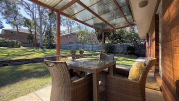 A covered patio with wicker furniture and a glass table, overlooking a backyard with a lawn and a wooden fence.