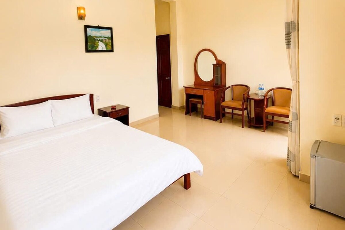 A hotel room with a large bed, a desk with two chairs, a small table, a mirror, a picture on the wall, and a radiator.