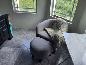 A room with a grey armchair, a matching ottoman, a wooden table, and a view through a window. - Executive One-Bedroom Apartment Boasting Natural Light and Lush Scenery! (Charlotte Amalie)
