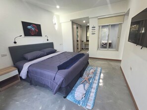 A bedroom with a bed, a bedside table, a wall-mounted light, a framed picture, a window with blinds, and a door leading to another room. - Spacious & Stylish 2-Bedroom Condo at Replay Residence – Bo Phut (Koh Samui)