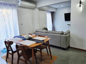 A modern living room with a dining area, grey sofa, and a flat-screen TV mounted on the wall. - Spacious & Stylish 2-Bedroom Condo at Replay Residence – Bo Phut (Koh Samui)