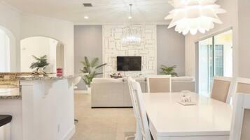 A modern living room with a sofa, a dining table with chairs, a TV, and a chandelier.