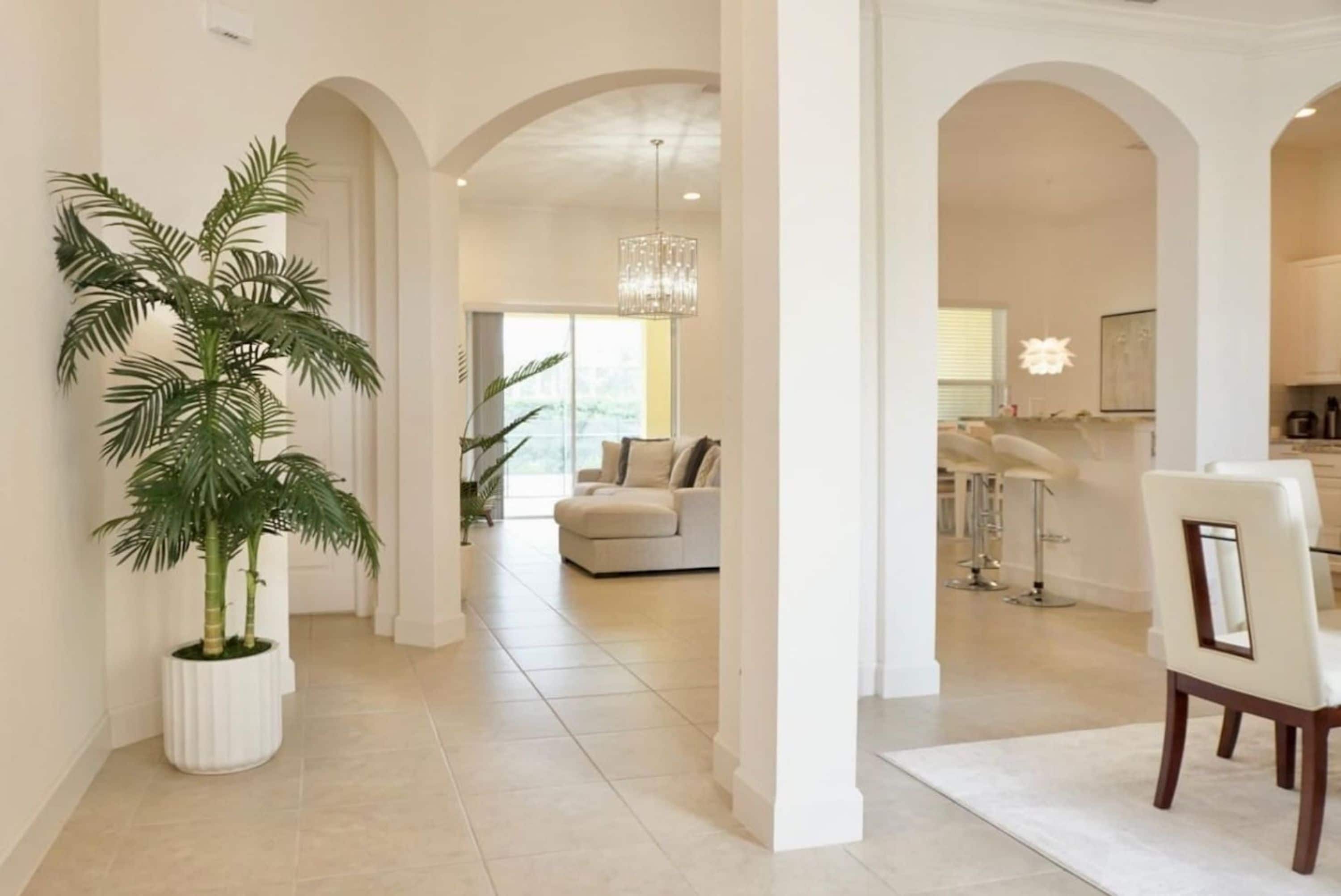 A spacious hallway with a large potted plant, a modern chandelier, and a view into a living area with a sofa and a dining area with bar stools.