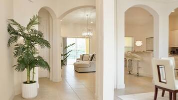 A spacious hallway with a large potted plant, a modern chandelier, and a view into a living area with a sofa and a dining area with bar stools.
