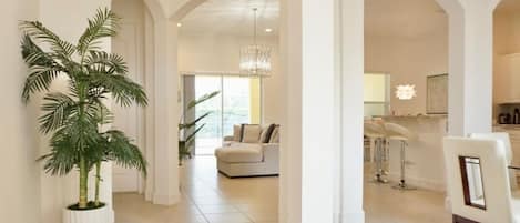 A spacious hallway with a large potted plant, a modern chandelier, and a view into a living area with a sofa and a dining area with bar stools.