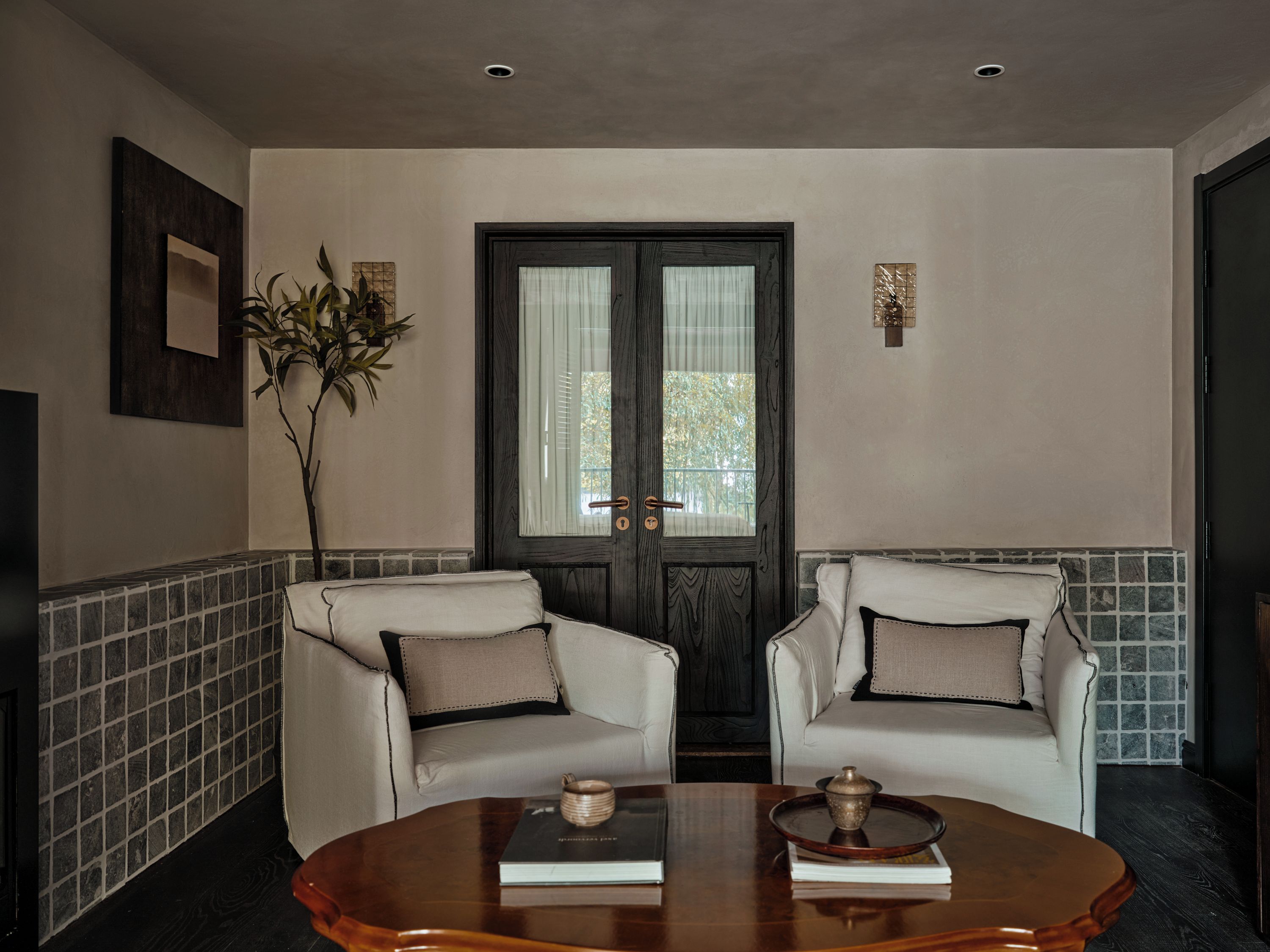 A room with two white armchairs, a wooden coffee table with books and a decorative item, a door with a window, and a wall-mounted picture.