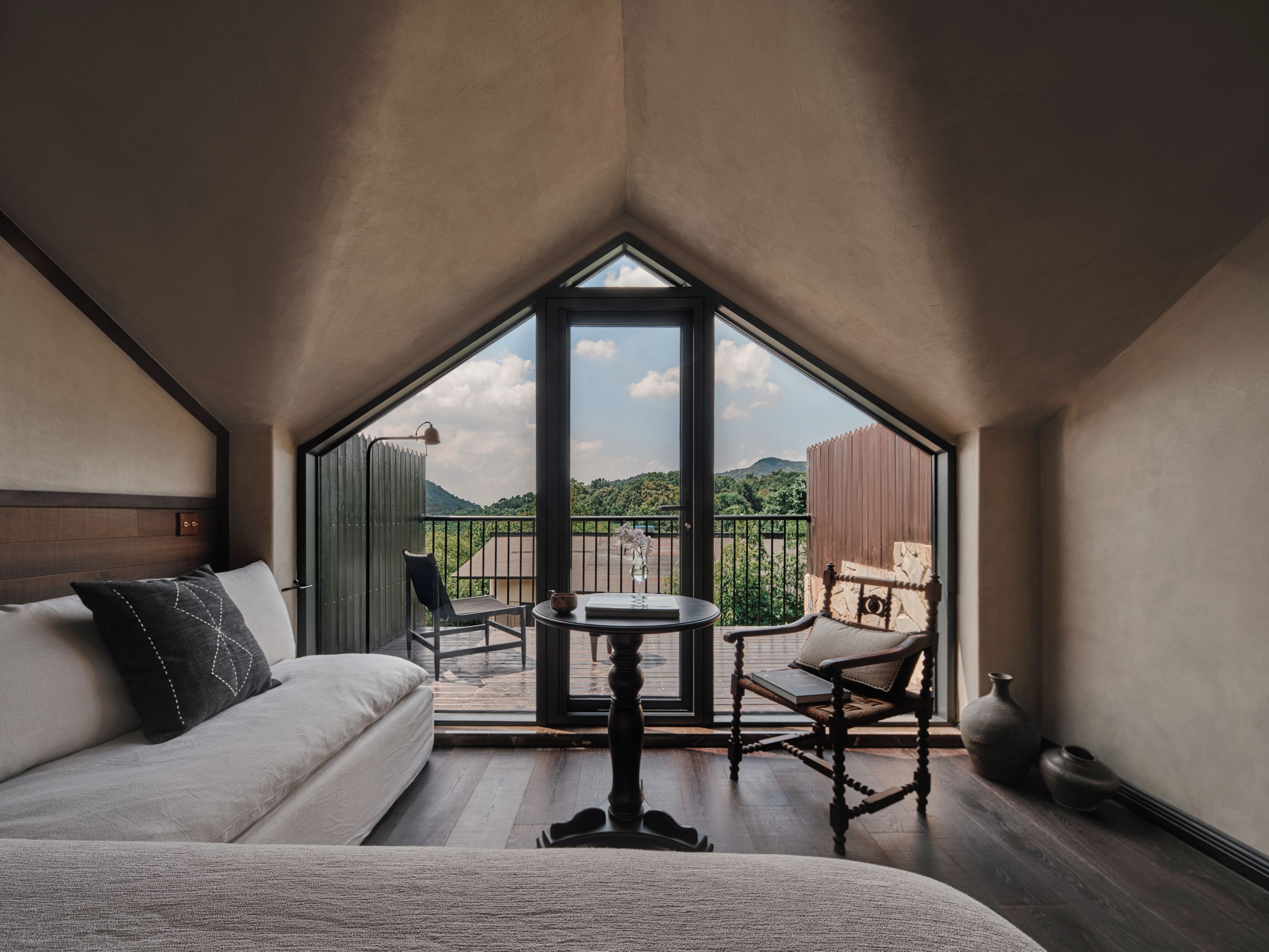 A room with a bed, a table, and a balcony with a view of greenery and mountains.