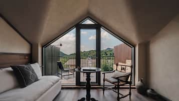 A room with a bed, a table, and a balcony with a view of greenery and mountains.