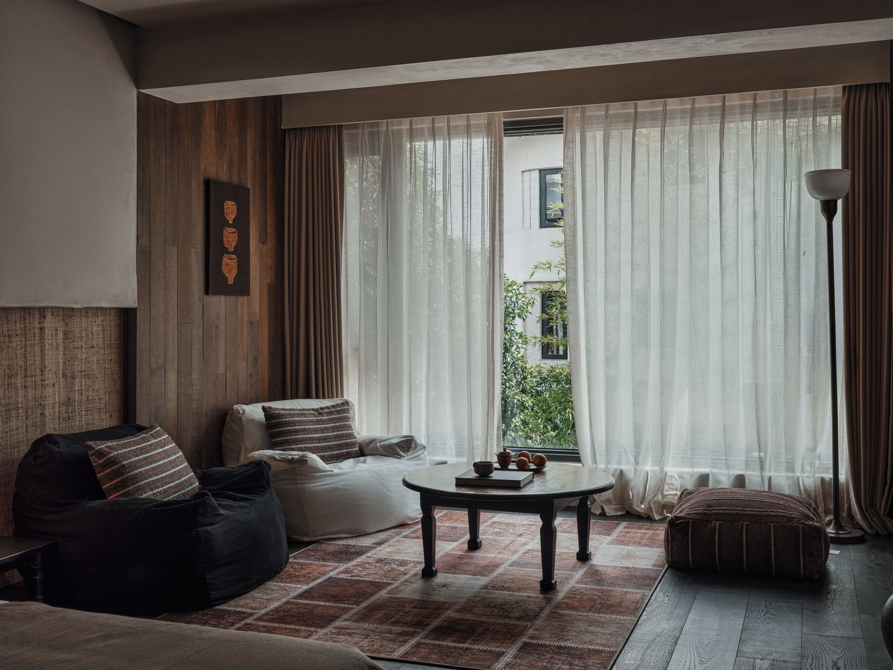A living room with a sofa, a coffee table, and a lamp. There is a window with curtains and a view of greenery outside.