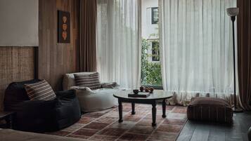 A living room with a sofa, a coffee table, and a lamp. There is a window with curtains and a view of greenery outside.