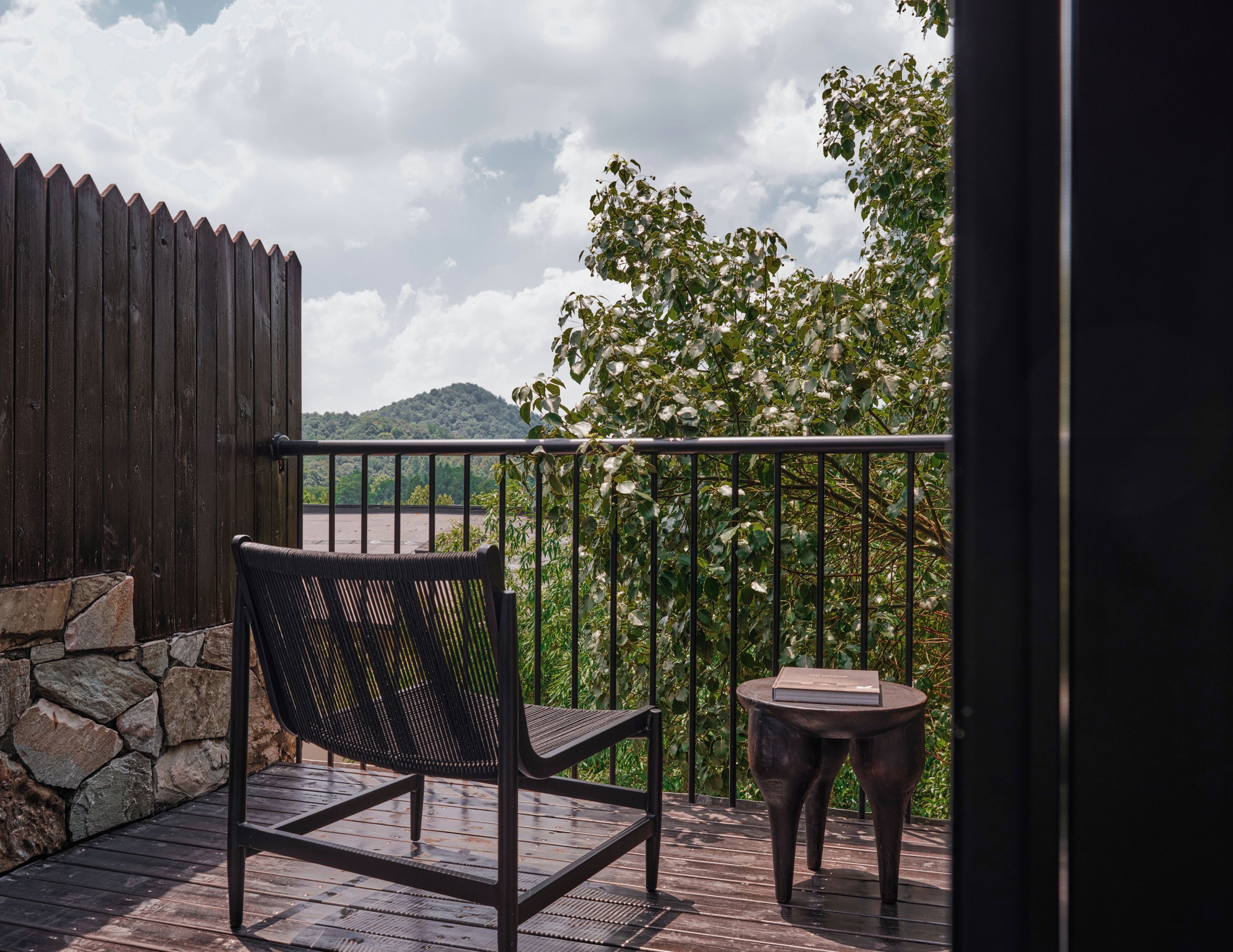 A wooden deck with a chair and a small table, a stone wall, and a view of a hill and trees.