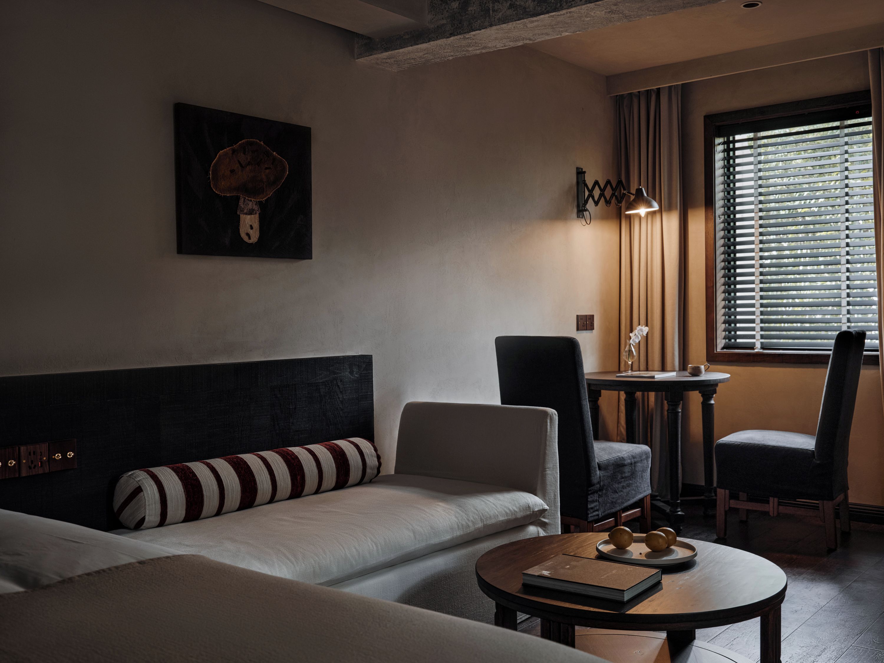 Ein modernes Hotelzimmer mit einer Sitzecke, zwei Stühlen, einem kleinen Tisch mit Buch und Dekoration, einer Lampe und einem Fenster mit Jalousien.