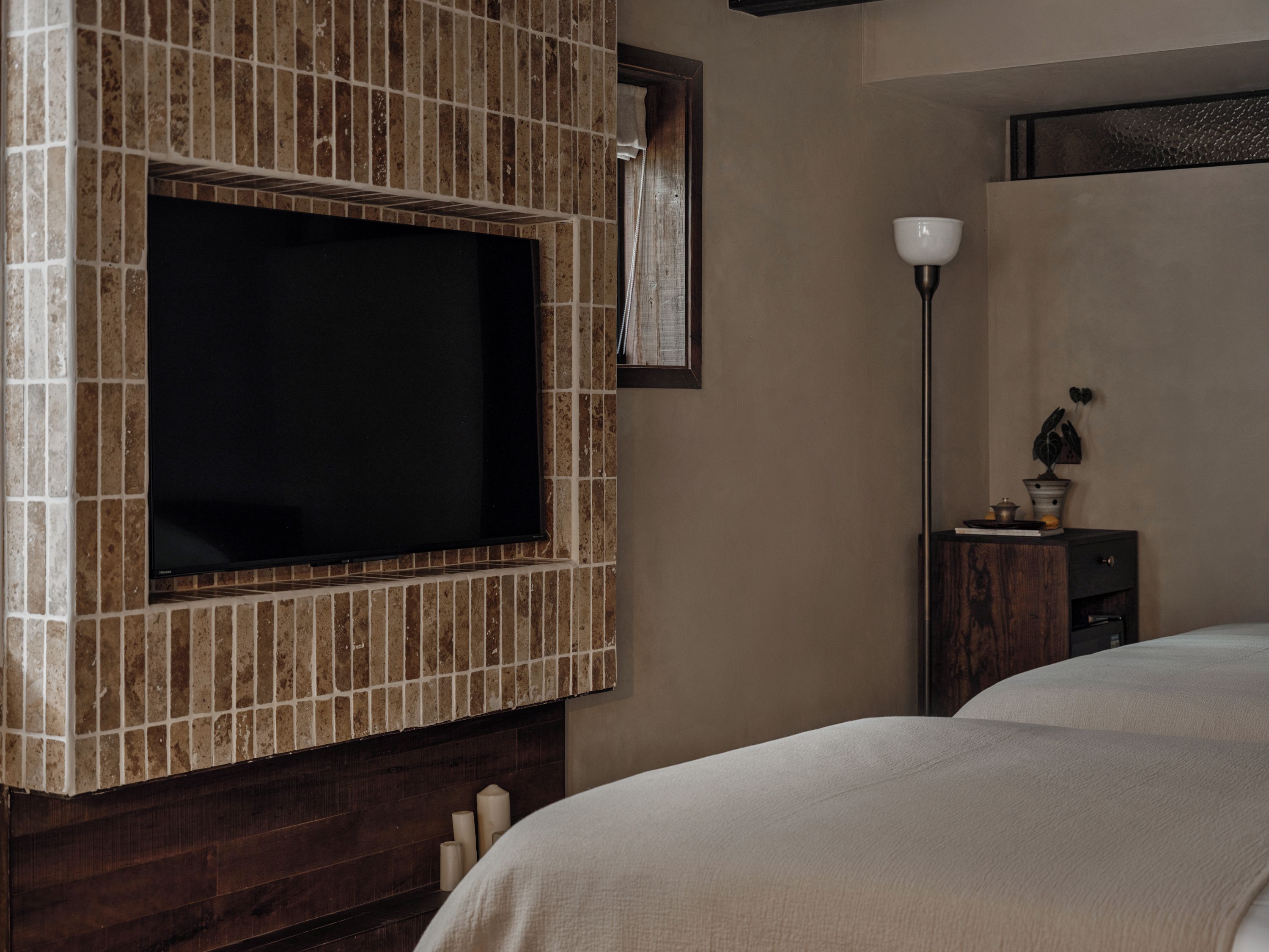 Habitación con una televisión instalada en una pared de azulejos, una mesita de noche con lámpara y una cama con cubierta blanca.
