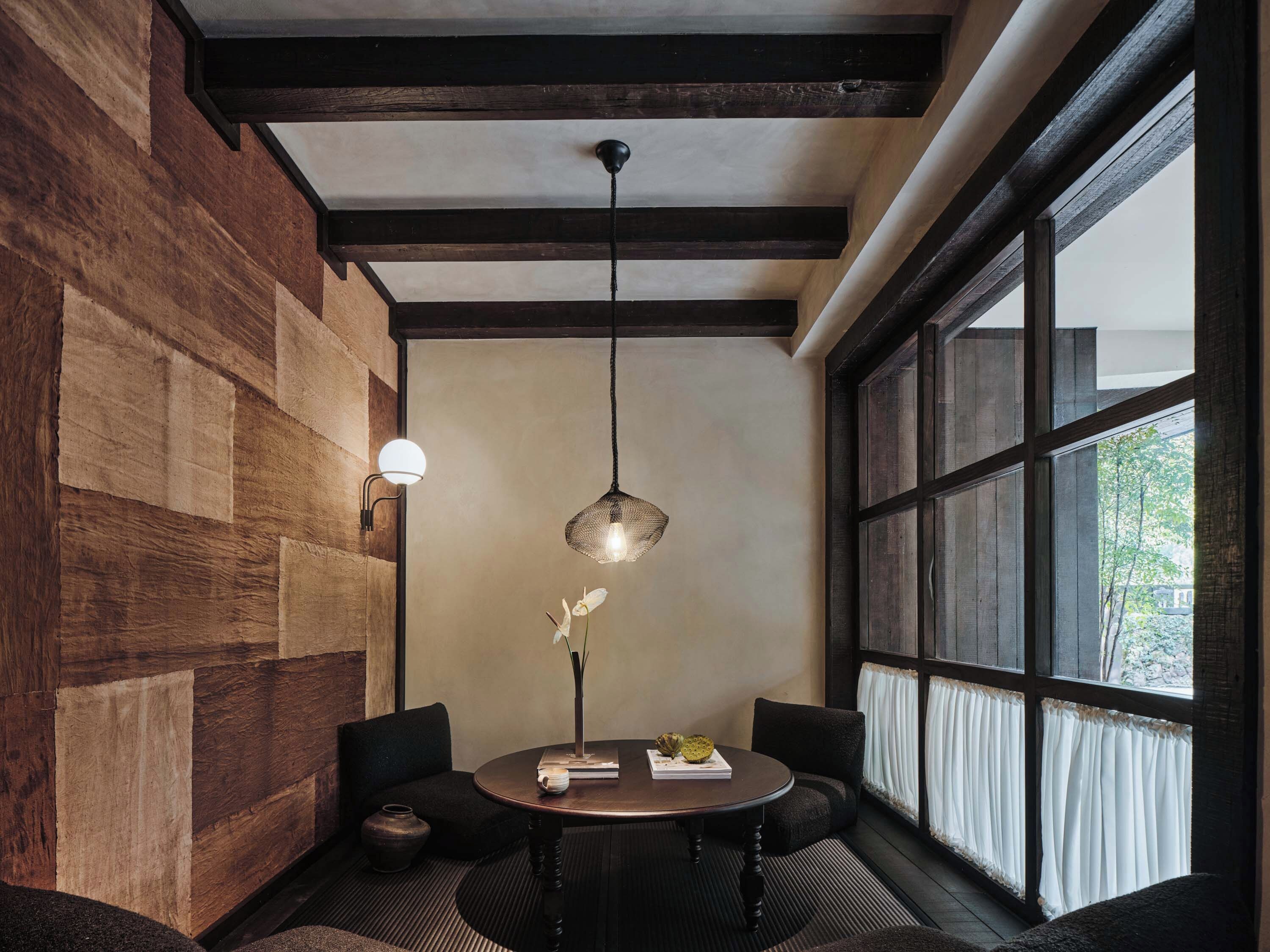 A room with a round table, two chairs, a hanging light fixture, and a large window with curtains.