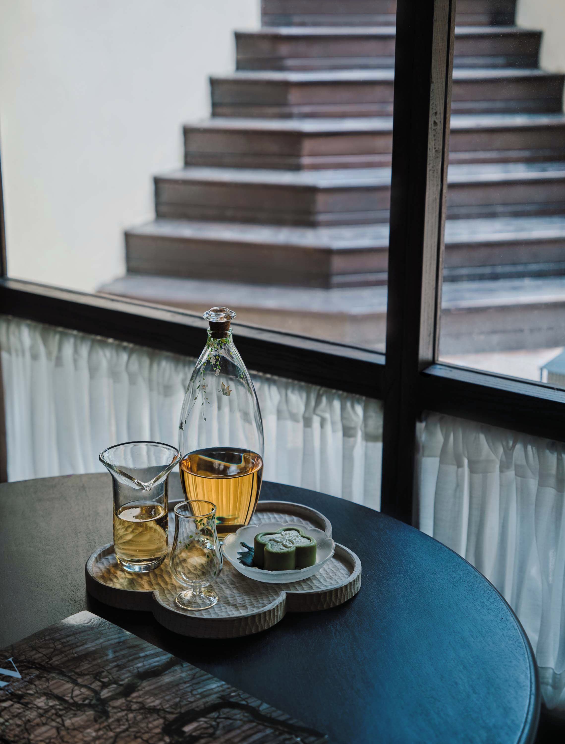 Una mesa con una botella, vasos y un plato con algo verde, frente a una ventana que da a una escalera.