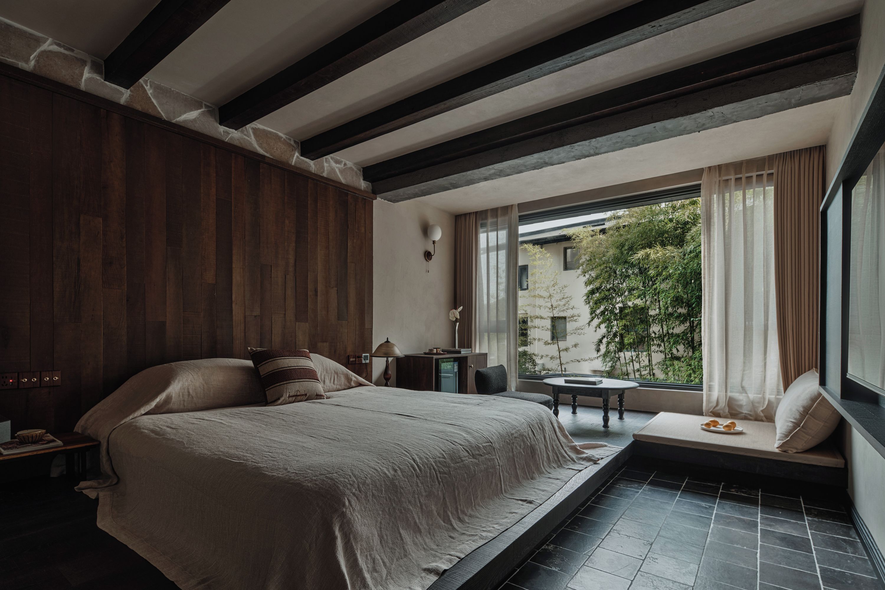 A bedroom with a large bed, a small table, and a view of greenery outside.