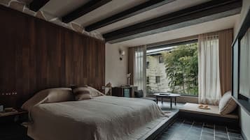 A bedroom with a large bed, a small table, and a view of greenery outside.
