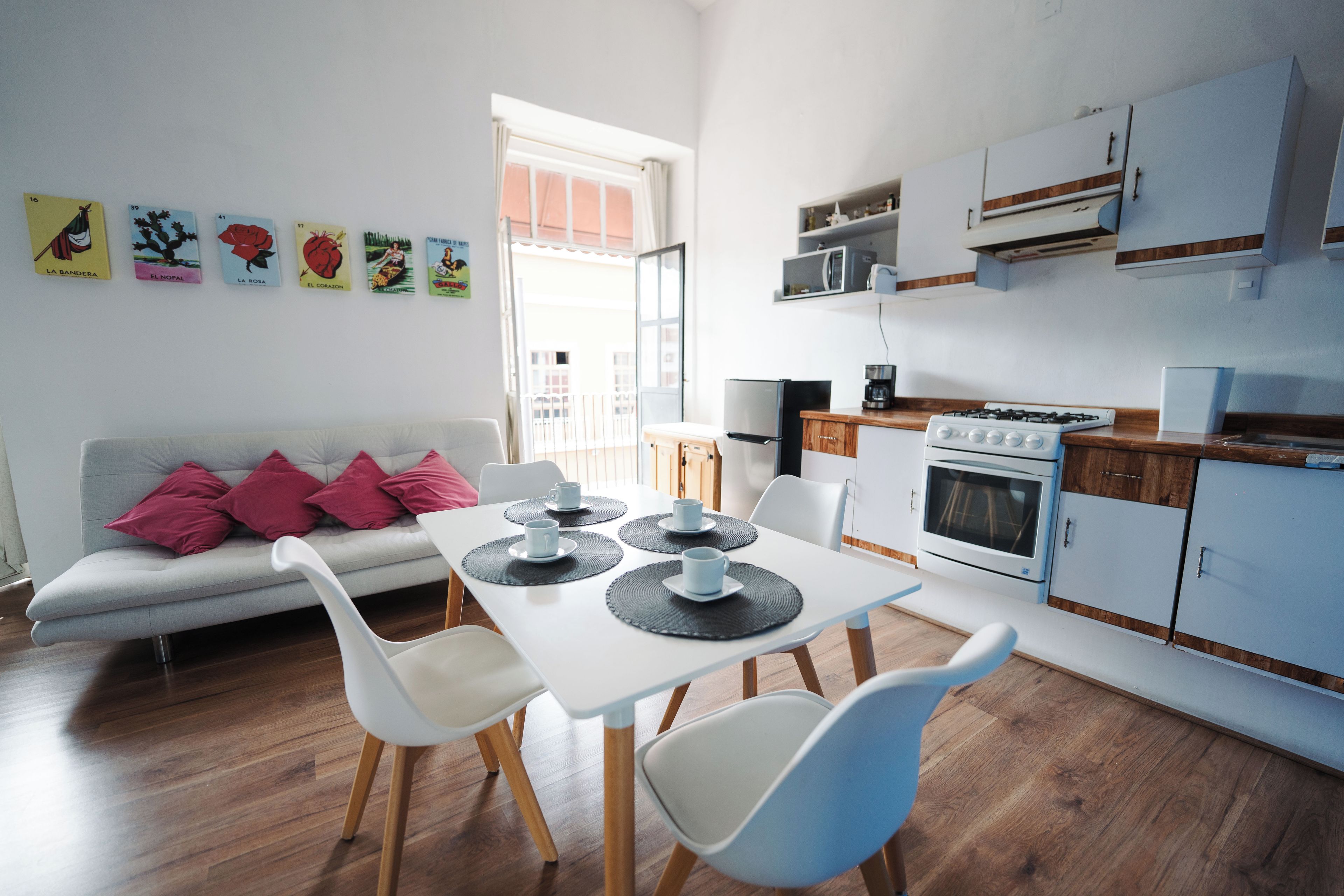 A modern kitchen with white cabinets, a stove, and a microwave. There is a dining table with chairs and a sofa with pink cushions.