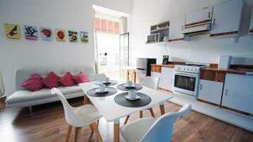 A modern kitchen with white cabinets, a stove, and a microwave. There is a dining table with chairs and a sofa with pink cushions.