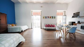 A spacious room with a wooden floor, a bed, a sofa, a dining table with chairs, and a kitchenette.