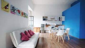 Um espaço de estar moderno com sofá branco, área de jantar com cadeiras brancas, cozinha com armários brancos e uma parede azul de destaque.