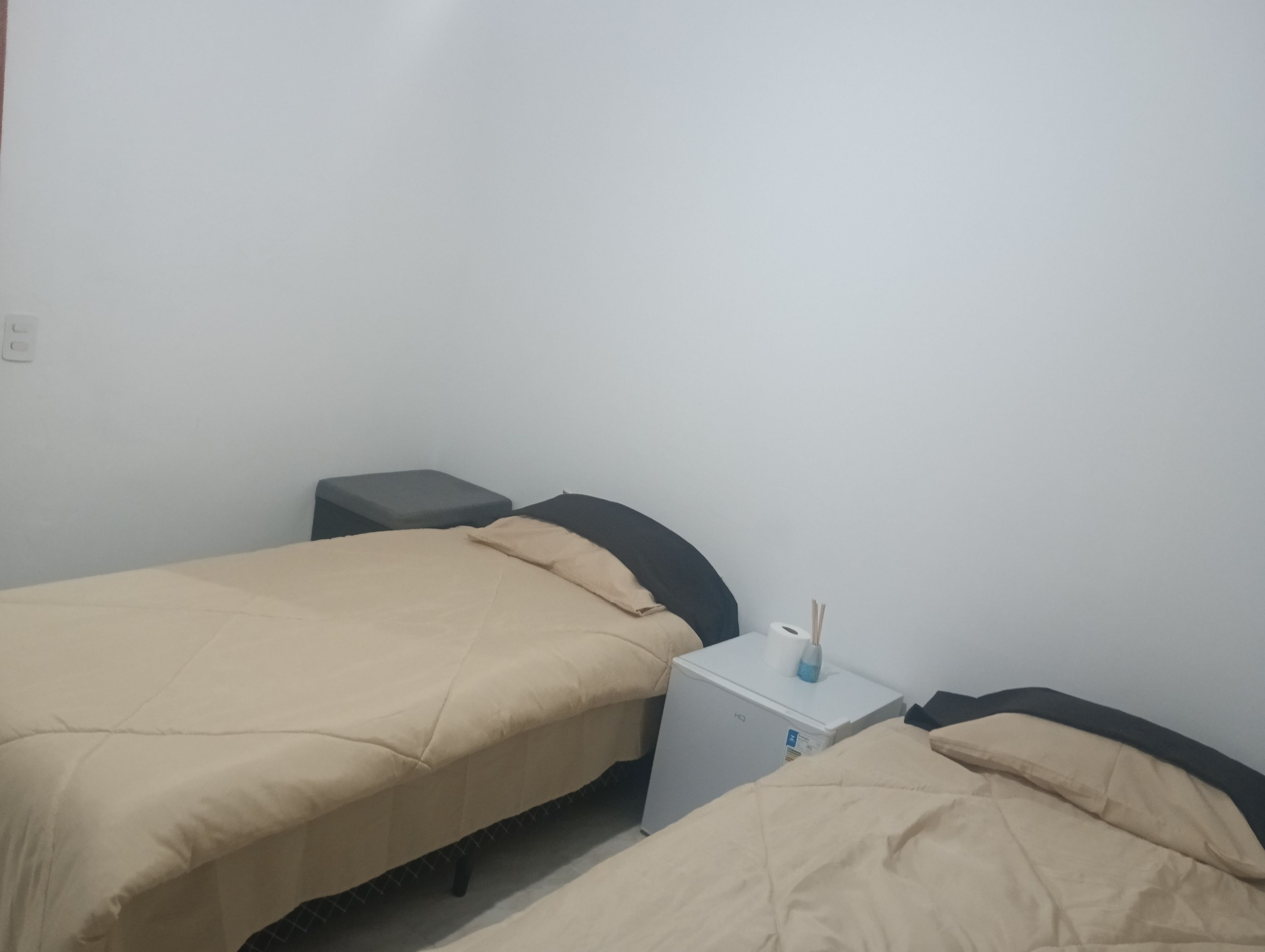 Ein kleines, schlichtes Zimmer mit zwei Betten, einem kleinen Tisch und einer weißen Wand.
